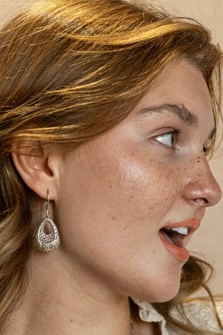 Side profile of a woman wearing detailed silver teardrop earrings with engraved patterns, shown against a soft neutral background.